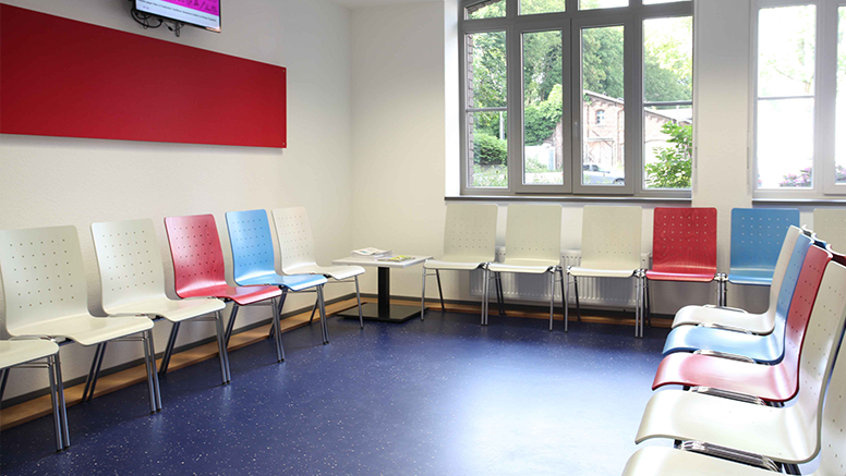 Sie sehen den Wartebereich des Jobcenters Wuppertal, Stühle nebeneinander gereiht mit Lehne zur Wand in rot, weiß und hellblau.