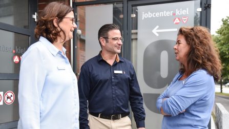 Zwei brünette Frauen in hellblauen Blusen und ein Mann mit Brille und blauem Hemd stehen vor der Tür des Jobcenters und unterhalten sich. (Bild anzeigen)