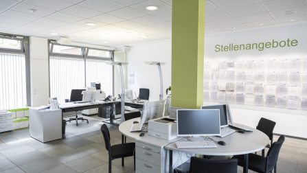 Ein heller Raum mit einer grünen Säule in der Mitte, mehreren Computerarbeitsplätzen und einer Wand, an der Stellenangebote aushängen. (Bild anzeigen)