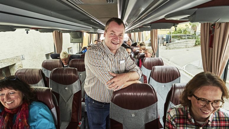 Peter Schadl trägt ein kariertes Hemd, steht im Gang eines Reisebusses und lacht.