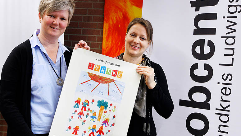 Barbara Schilling und Sonja Ohren halten ein Plakat hoch, auf dem "Landesprogramm STÄRKE" und ein von Kindern gemaltes buntes Bild gedruckt sind. 