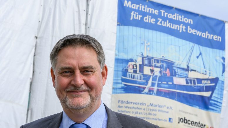 Martin Greiner steht vor einer weißen Plane, darauf ein kleineres Plakat des Jobcenters mit Bild eines Schiffs unter der Überschrift "Maritime Tradition für die Zukunft bewahren".