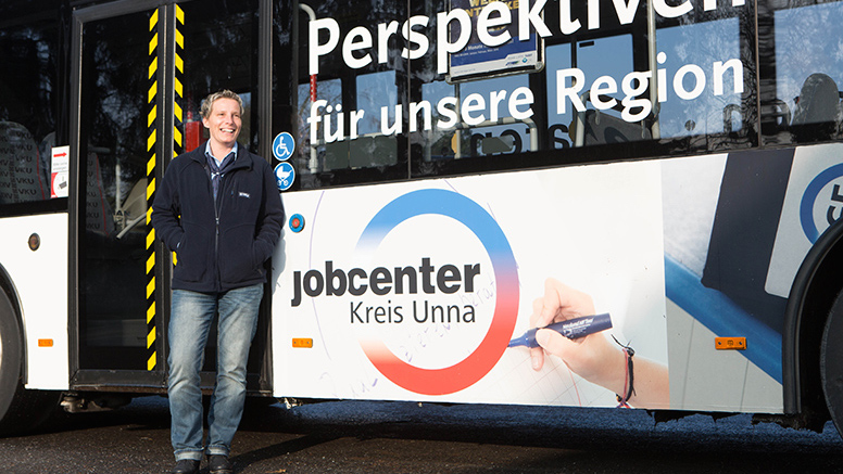 Heike Sitz hat kurze blonde Haare und steht in Arbeitskleidung vor einem Linienbus. Auf ihm steht "Perspektiven für unsere Region. Jobcenter Kreis Unna".