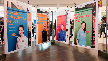 Vier großflächige Plakate hängen an Bauzaun, auf ihnen vier Portraits von jungen Menschen.OeffnetSeiteU25-Haus Mülheim