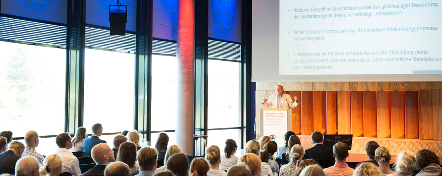 Prof. Dr. Claus Reis steht auf einer Bühne vor Publikum, im Hintergrund eine Präsentation zum "Produktionsnetzwerk".