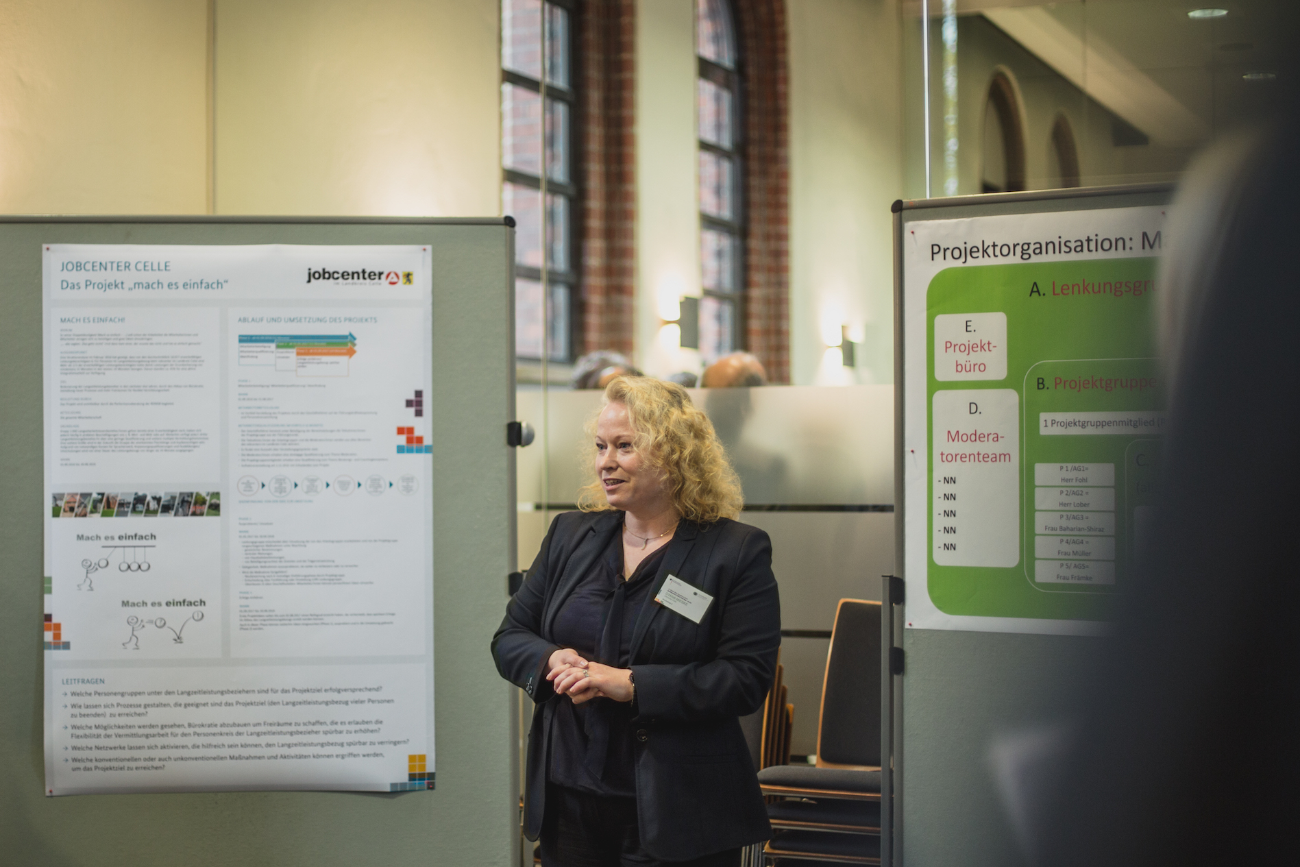 Eine Frau mit blonden Locken steht vor einem Plakat des Jobcenters Celle und präsentiert (Bild anzeigen)