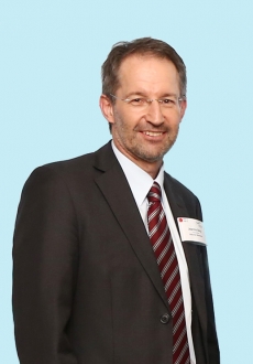 Portrait von Joachim Burg. Er hat kurze braun-graue Haare, trägt eine randlose Brille und einen schwarzen Anzug. 