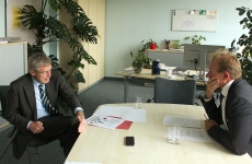 Dr. Ulrich Gawellek und der Interviewer sitzen gegenüber an einem Tisch. Beide haben kurze Haare und tragen einen schwarzen Anzug. 