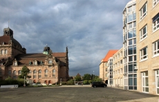 Blick auf einen asphaltierten Platz. Links die barocke, braun-beige Staatsoper und rechts das moderne, helle Jobcentergebäude.