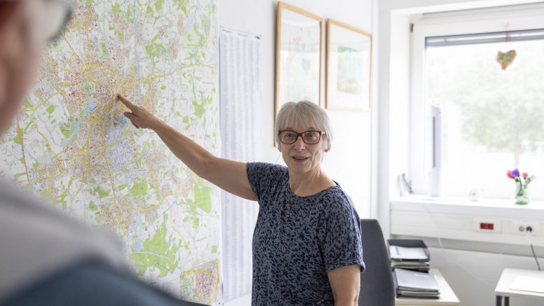 Eine Frau mit Brille deutet auf einen großen Stadtplan von Münster, der an einer Bürowand hängt.