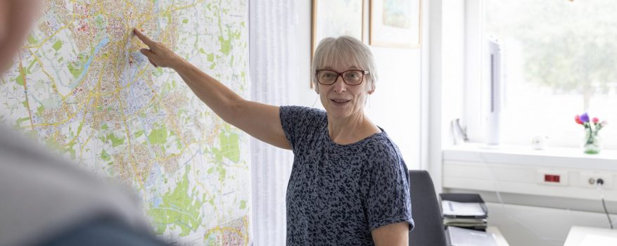 Eine Frau mit Brille deutet auf einen großen Stadtplan von Münster, der an einer Bürowand hängt.
