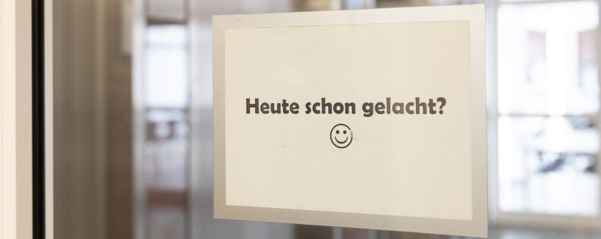 An einer Glastür hängt ein Schild mit der Aufschrift "Heute schon gelacht?" und einem Smiley darunter.