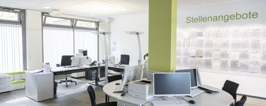 Ein heller Raum mit einer grünen Säule in der Mitte, mehreren Computerarbeitsplätzen und einer Wand, an der Stellenangebote aushängen.