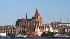 Sicht auf eine emporragende gotische Kirche sowie Häuser in hanseatischer Bauweise am Meer.Öffnet Seite: Jobcenter-Porträt Rostock