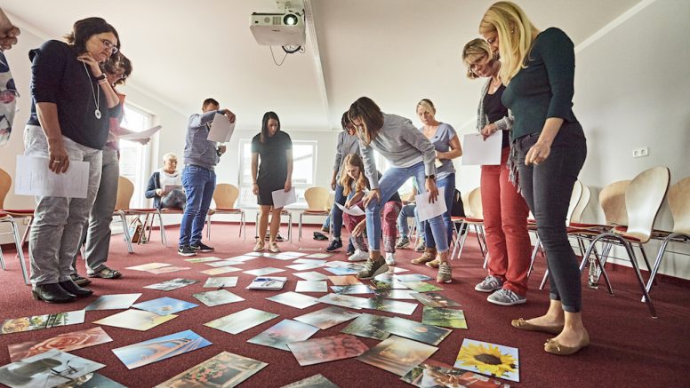 Jobcenter-Beschäftigte stehen bei einem Workshop im Kreis. Auf dem Boden in der Mitte liegen Fotos.