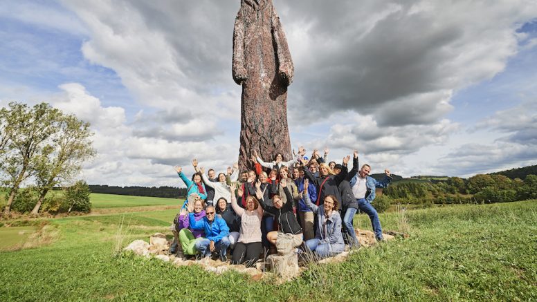 Gruppenfoto vor einer großen braunen Statue, drumherum grüne Wiesen. 