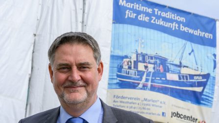 Martin Greiner steht vor einer weißen Plane, darauf ein kleineres Plakat des Jobcenters mit Bild eines Schiffs unter der Überschrift 