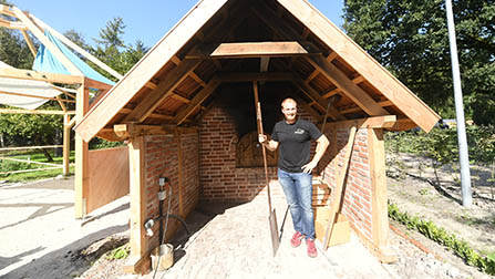 Ein Mann steht unter dem Backhause, das Backsteinwänden und Holzdach hat. (Bild anzeigen)
