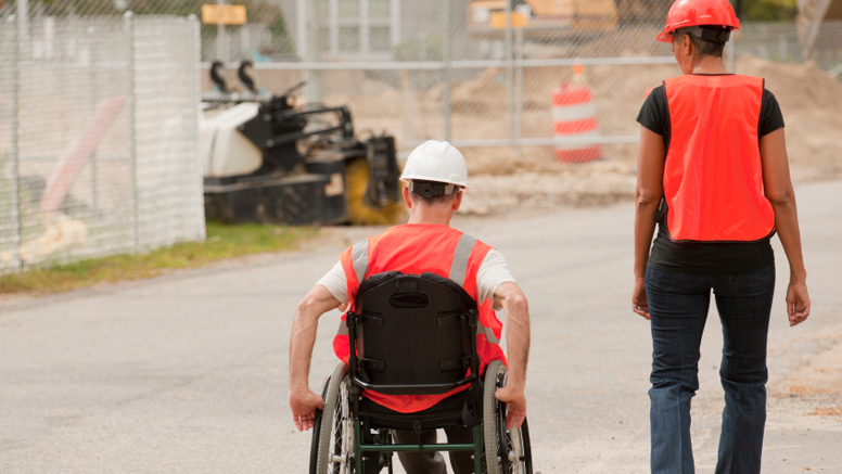 Ein Mann im Rollstuhl trägt eine Warnweste und einen Bauhhelm.