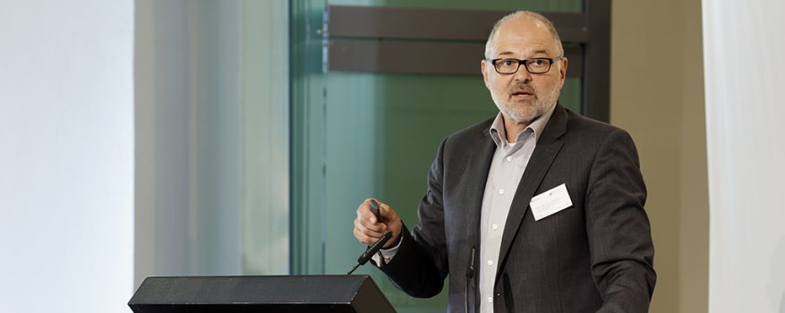 Porträtfoto Dr. Kupka. Er steht hinter einem Pult und trägt vor.